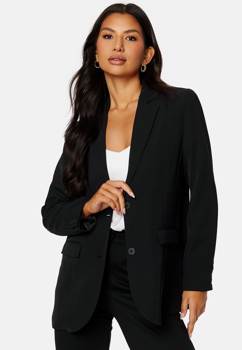 Czarna marynarka o dopasowanym kroju, z klapami, z pojedynczym guzikiem i dwiema przednimi kieszeniami. Noszona z białą bluzką pod spodem.