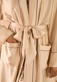 Robe in satin beige con vita cintata, caratterizzato da piping viola lungo i bordi e due tasche frontali. Texture liscia e design aperto.