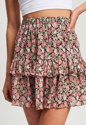 Kobieta nosząca warstwową kwiecistą spódnicę z różowymi, białymi i zielonymi kwiatami na czarnym tle oraz elastycznym pasem w talii.