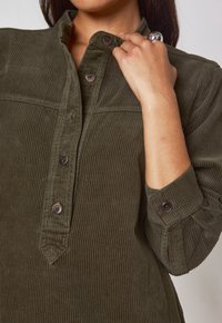 Camicia in corda verde oliva con colletto, chiusura a bottoni e maniche arrotolate. Tessuto testurizzato con un sottile motivo a righe verticali.