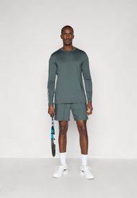 Σετ αθλητικής ένδυσης που περιλαμβάνει μακρυμάνικο σκούρο πράσινο μπλουζάκι και ταιριαστά σορτσάκια. Κρατάει μια ρακέτα τένις ενώ φοράει λευκά αθλητικά παπούτσια και κάλτσες.