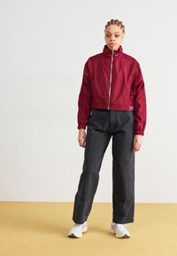 Maroon cropped jacket with a high collar and front zipper, paired with loose black pants and white sneakers on a light yellow background.