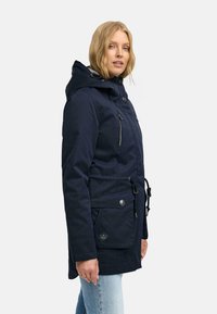 Námornícky modrý parka s kapucňou, so zipsovými a patentkovými vreckami, nastaviteľným sťahovaním v páse a hladkou textúrou; nosená s svetlomodrými džínsami.