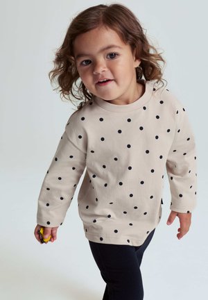 Beige Langarmhemd mit marineblauen Punkten, runder Ausschnitt und weichem Stoff, kombiniert mit schwarzen Leggings. Kind, das ein gelbes Spielzeug hält.