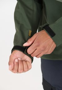 Veste imperméable verte avec réglage des poignets en velcro. Détail de logo visible en jaune sur la manche. Texture de tissu lisse.