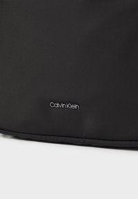 Borsa nera in nylon con superficie texturizzata, caratterizzata da un logo Calvin Klein discreto in bianco. Design elegante e minimalista senza ulteriori accentuazioni.