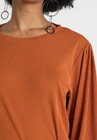 Rostfärgad topp med rund halsringning, mjuk textur och skrynkliga detaljer på ärmarna. Har stora cirkulära örhängen.