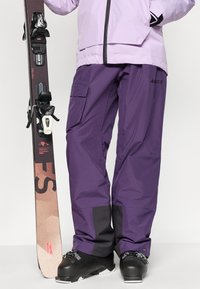 Pantalones de esquí en tela impermeable morada, con bolsillos cargo, acentos negros y un ajuste holgado. Se sostiene un esquí con fijaciones al lado.