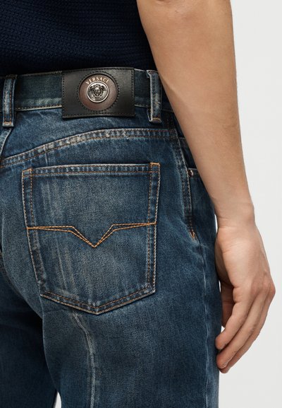 Buzunar Jean cu cusături portocalii, etichetă din piele Versace și prim-plan cu brațul drept și mâna unei persoane pe un fundal simplu.