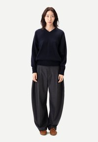 Marinblå ulltröja med V-ringning och ribbad fåll, kombinerad med grå vidbyxade byxor och bruna flätade skor. Enfärgad bakgrund.