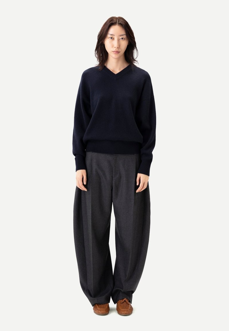 Marinblå ulltröja med V-ringning och ribbad fåll, kombinerad med grå vidbyxade byxor och bruna flätade skor. Enfärgad bakgrund.