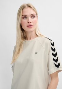 T-shirt couleur beige clair à manches courtes avec un col rond, avec des accents de motif en chevron noir sur les manches et un petit logo d'abeille brodé.