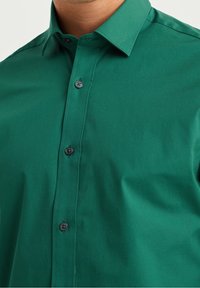 Grön knapp-up skjorta gjord av slät tyg, med en klassisk krage och svarta knappar. Närbild som framhäver texturen och färgen.