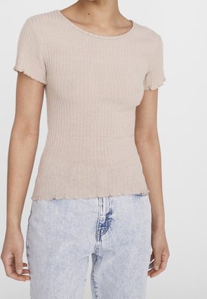 Frau trägt ein figurbetontes, beige geripptes Kurzarmoberteil mit welligem Saum und hellblaue, hoch taillierte Jeans, gezeigt vom Hals bis zu den Hüften.