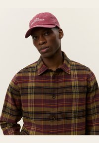 Gorra marrón con el texto "Lower West Side", combinada con una camisa de cuadros en tonos tierra de olivo, burdeos y mostaza. Diseño con botones y cuello.