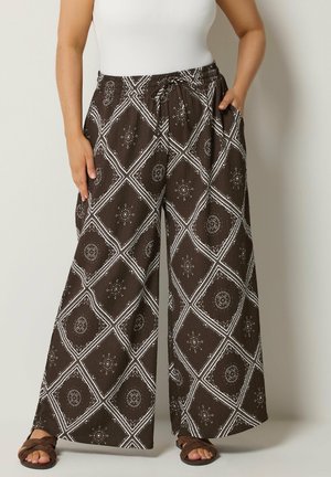 Pantalon large marron avec des motifs géométriques et circulaires blancs, porté avec des sandales et un haut sans manches blanc.