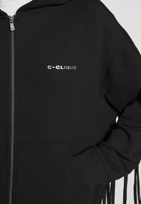 Sweat à capuche noir zippé avec poches avant et texte blanc "c-CLIQUE" sur la poitrine, porté par une personne avec la main gauche dans la poche.