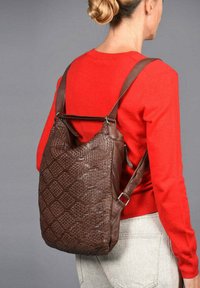 Bruine geweven leren rugzak met een gestructureerde vorm, verstelbare bandjes en een gladde afwerking, gedragen op de rug tegen een rode trui.