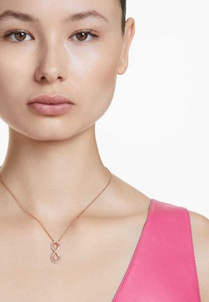 Halskette aus Roségold mit einem Unendlichkeit-Anhänger, verziert mit klaren Steinen, getragen über einem pinken Oberteil mit tiefem Ausschnitt. Glatte Hauttextur.