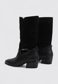 Botas negras hasta la rodilla con un tacón cubierto, que cuentan con un cuero texturizado brillante y una parte superior de gamuza suave. Incluyen un detalle decorativo de hebilla.