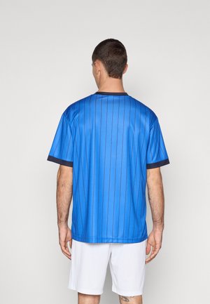 Maillot bleu avec des rayures verticales bleu marine, manches courtes et col rond bleu marine, en tissu synthétique lisse, porté avec un short blanc.