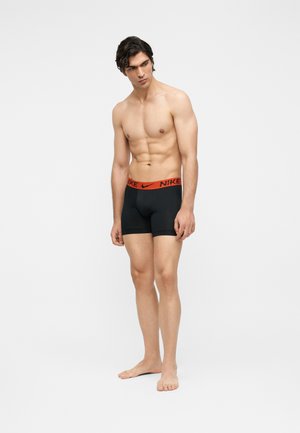 Homme athlétique debout pieds nus, portant un boxer Nike noir avec une ceinture rouge, regardant vers le bas et légèrement sur le côté sur un fond uni.
