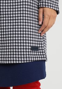 Ruka odpočívající na kabátu s černobílým vzorem houndstooth, s černou etiketou, vrstveném přes námořnické a červené oblečení.