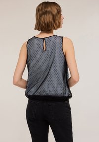 Transparante zwarte mouwloze top met een gestructureerd ontwerp, voorzien van een sleutelgat-detail op de rug, gedragen met zwarte jeans. Subtiele verticale patronen doorheen.