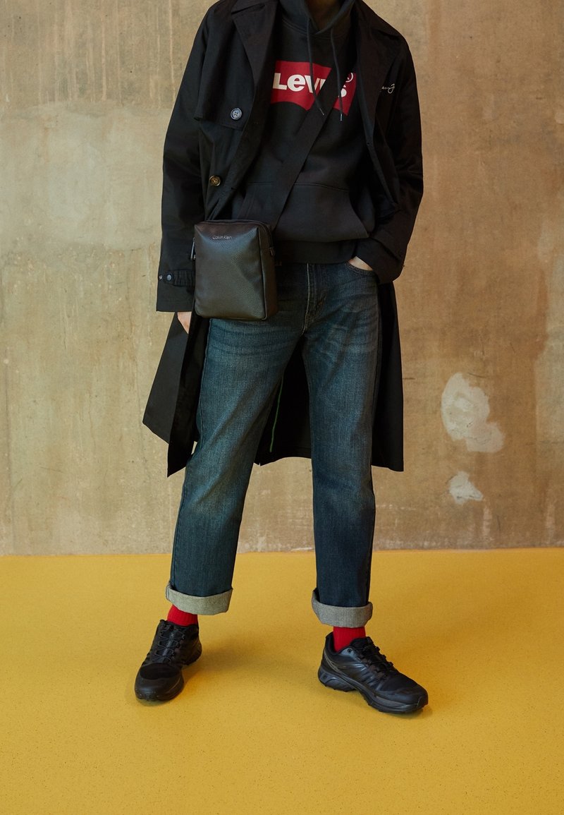 Abrigo negro sobre una sudadera gris con un logo rojo de Levi's, jeans azules con puños, calcetines rojos y zapatillas negras. Pequeño bolso negro cruzado.