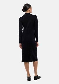 Robe noire à côtes avec des manches longues, longueur genoux. Présente une fente au dos et un design minimaliste. Portée avec des chaussures noires.