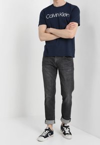 Man som bär en marinblå Calvin Klein t-shirt, rullade mörkgrå jeans och svarta Converse sneakers, står med armarna i kors.