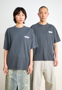 Gray oversized t-shirts with white printed text, featuring short sleeves and crew neck. Model wears a patterned skirt and comfortable trousers.
