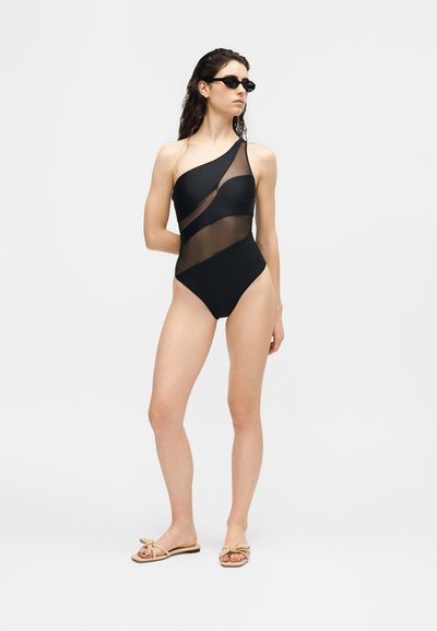 Γυναίκα που φοράει μαύρο ολόσωμο μαγιό με έναν ώμο και διαφανείς διαγώνιες ταινίες, μαύρα γυαλιά ηλίου και μπεζ σανδάλια, στέκεται μπροστά σε απλό φόντο.