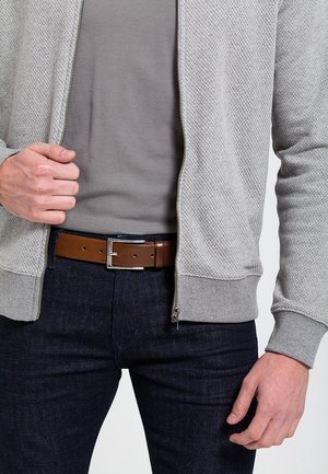 Mężczyzna ubrany w jasnoszary sweter na suwak, szarą koszulę, ciemne dżinsy i brązowy skórzany pasek z srebrną klamrą, ręka regulująca suwak swetra.