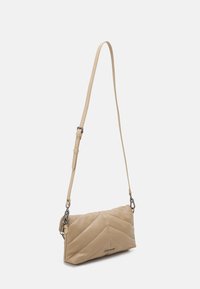 Beżowa prostokątna torebka na ramię z pikowanym wzorem w jodełkę, regulowanym paskiem i metalową plakietką z logo z przodu na białym tle.