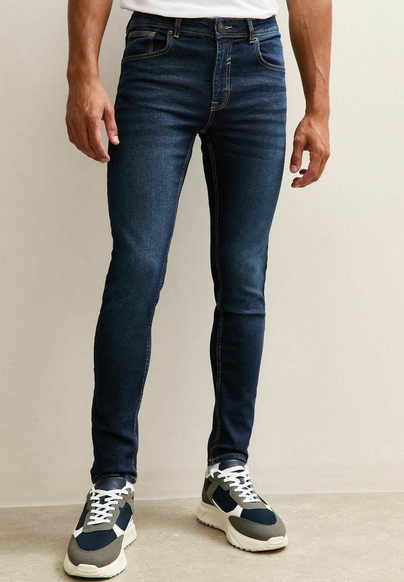 Man som bär mörkblå slim fit-jeans och svarta, grå och vita sneakers står på ett beige golv mot en ljus vägg.