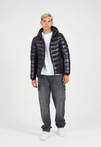 Črna puhovka s kapuco, z bleščečo teksturo in kvadratnim vzorcem. Obuta je nad belo majico, s sivimi cargo hlačami in belimi čevlji.