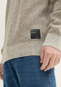 Hellbrauner Strickpullover mit strukturiertem Muster, ausgestattet mit einem schwarzen Markenetikett an der Seite, kombiniert mit blauen Denim-Jeans.