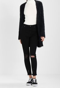 Svarta ripped skinny jeans, en fluffig svart cardigan, en vit ribbad turtleneck och svarta sneakers med vita snören på en neutral bakgrund.