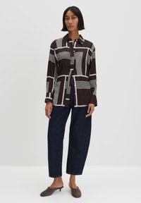 Camicia marrone con motivi geometrici bianchi, design con bottoni, maniche lunghe, colletto, abbinata a pantaloni di jeans scuri e mule marroni.