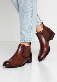 Botas de cuero marrón hasta el tobillo con paneles laterales elásticos, tacón bajo y textura suave, combinadas con jeans desgastados de color azul claro.