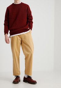 Punainen neulepaita, vaaleat housut, ruskeat kengät, valkoiset sukat. Neulepaidassa on teksturoitu pinta, jossa on resorit hihoissa ja helmassa. Housut ovat löysät.