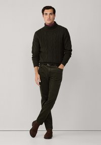 Pull en torsades vert foncé à col roulé, pantalon en velours côtelé vert foncé à coupe ajustée, et bottines en suède marron, créant un look coordonné.