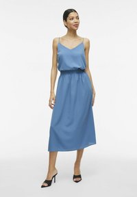 Robe maxi sans manches bleue avec un décolleté en V, taille froncée et tissu fluide. Le modèle porte des mules ouvertes noires. Texture lisse, design décontracté.