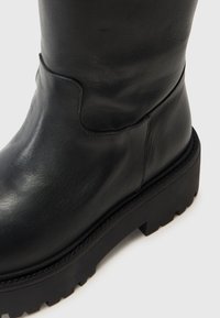Botine negre din piele, cu o textură netedă, vârf rotunjit și talpă groasă din cauciuc negru cu un model de tip lug. Design minimalist.