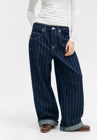 Jean en denim bleu foncé avec des rayures blanches. Design à jambes larges, grandes poches et revers. Fermeture bouton à la taille.