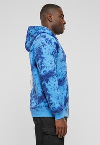 Hoodie bleu tie-dye avec une capuche à cordon de serrage, poche kangourou et poignets côtelés. Le matériau présente une finition douce et texturée.