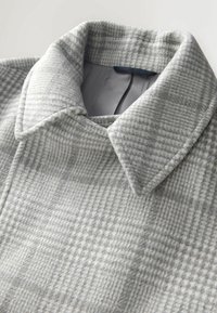 Harmaa ruutukuvioinen villakangas, jossa on pehmeä rakenne; klassinen kaulus ja jämäkkä kaulusrakenne; hienovaraisia kuviointivaihteluita.