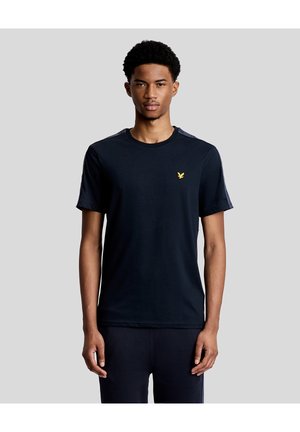 Mand iført en sort T-shirt med korte ærmer og et lille gul fuglelogo på venstre bryst, stående mod en ensfarvet grå baggrund.