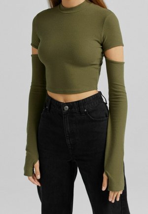 Kobieta w oliwkowozielonym skróconym topie z odłączanymi długimi rękawami i wysokim stanem czarnych dżinsów na jednolitym tle.